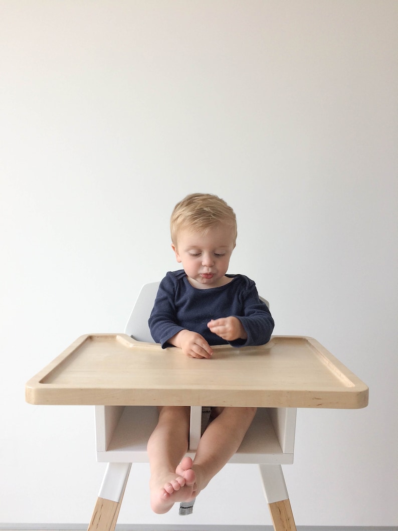Op de afbeelding: Een houten kinderstoel met een lichtgekleurde lade en witte poten. De kinderstoel heeft een natuurlijke houten afwerking en een eenvoudig, modern ontwerp. Het kind draagt een marineblauw shirt met lange mouwen.