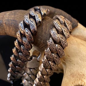 Op de afbeelding: Twee zilverkleurige armbanden met een ingewikkeld, zich herhalend patroon. De armbanden worden getoond op een botkleurig oppervlak, met een donkere achtergrond. Het ontwerp heeft een reeks gestileerde, gebogen vormen.