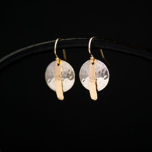 May include: A pair of silver-colored disc earrings with a hammered texture, each featuring a vertical gold-colored bar. The earrings have gold-colored ear wires and hang against a black background. The jewelry is simple and elegant.