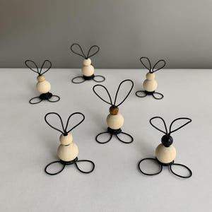 May include: Six decorative bunny ornaments. Each has a wooden bead body, wire ears, and a wire base. Some have a dark bead for the head, others a natural wood bead or a black bead. Ideal for Easter.