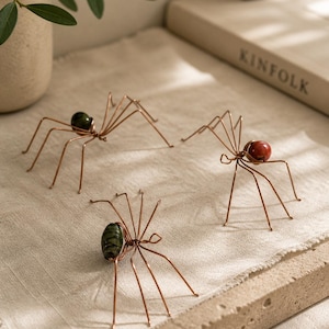 Puede incluir: Tres arañas decorativas de alambre de cobre con cuerpos de piedras preciosas. Una araña tiene un cuerpo verde, otra un cuerpo rojo y la tercera un cuerpo verde y negro. Las arañas se exhiben sobre una superficie beige.