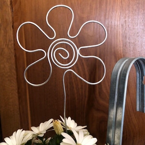 Könnte beinhalten: Eine silberne Drahtblume mit sechs Blütenblättern und einer spiralförmigen Mitte. Die Blume steht in einem Topf mit weißen Gänseblümchen.