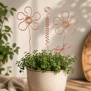 Può includere: Un piccolo vaso di ceramica macchiata pieno di piante verdi, adornato con tre sculture floreali in filo di rame. I fiori hanno un centro a spirale e un design a petali. Il vaso è su una superficie di legno.
