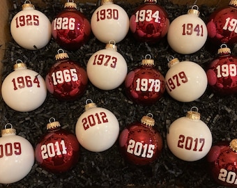 Könnte beinhalten: Eine Sammlung von 14 roten und weißen Weihnachtskugeln mit goldenen Akzenten. Jede Kugel zeigt ein weißes Jahr in rot gedruckt, von 1925 bis 2020.