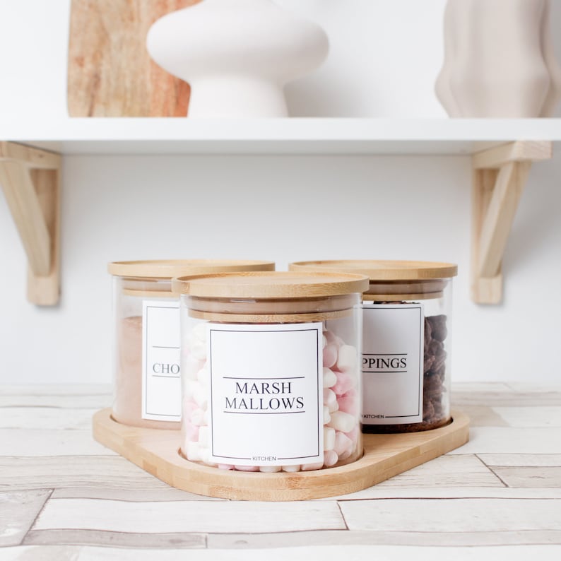 May include: Three clear glass jars with bamboo lids sit on a wooden tray. The jars are labeled "CHO", "MARSHMALLOWS", and "TOPPINGS". The marshmallows are pink and white. The jars are on a light-colored surface.
