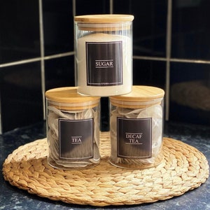 May include: Three glass kitchen canisters with bamboo lids, labeled "SUGAR", "TEA", and "DECAF TEA". The canisters sit on a woven placemat. The background features black tile.