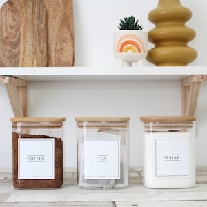 May include: Three clear glass kitchen canisters with bamboo lids, labeled "COFFEE," "TEA," and "SUGAR." The coffee canister is filled with brown coffee beans, the tea canister with tea bags, and the sugar canister with white sugar. The labels are white with black text.