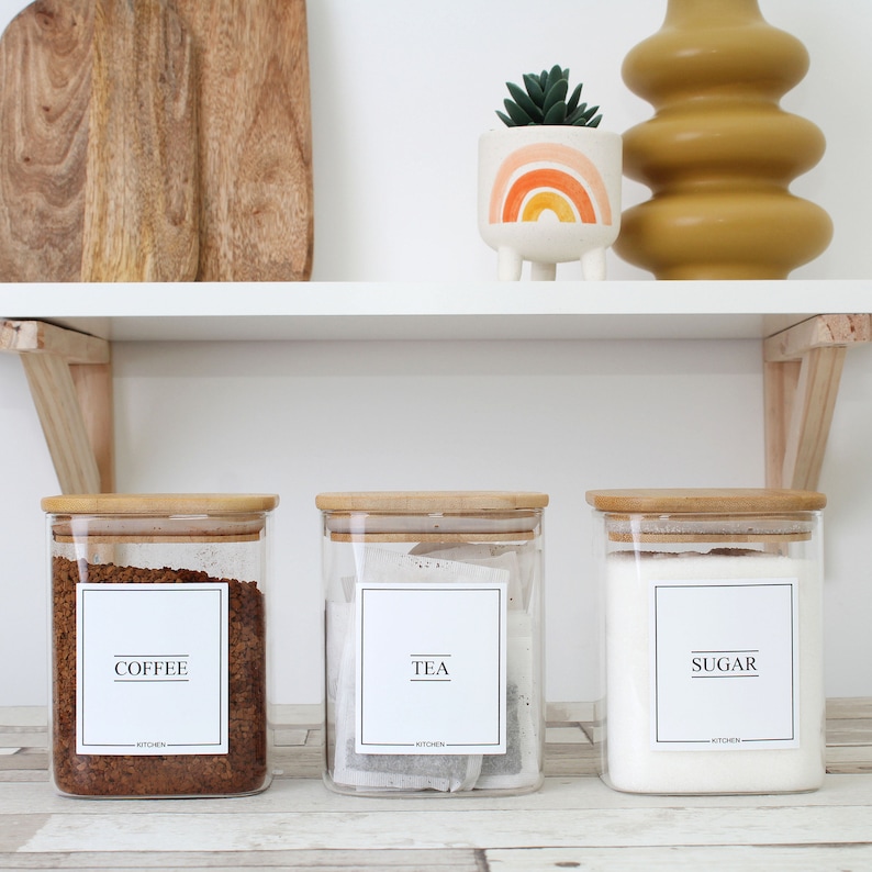 May include: Three clear glass kitchen canisters with bamboo lids, labeled "COFFEE," "TEA," and "SUGAR." The coffee canister is filled with brown coffee beans, the tea canister with tea bags, and the sugar canister with white sugar. The labels are white with black text.