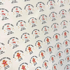 Op de afbeelding: Een vel kerststickers. De stickers tonen een sneeuwpop met een rode hoed en sjaal, en een peperkoekmannetje met een rode sjaal. Tekst bevat "Vrolijk Kerstfeest" en namen als "Phoebe" en "James".