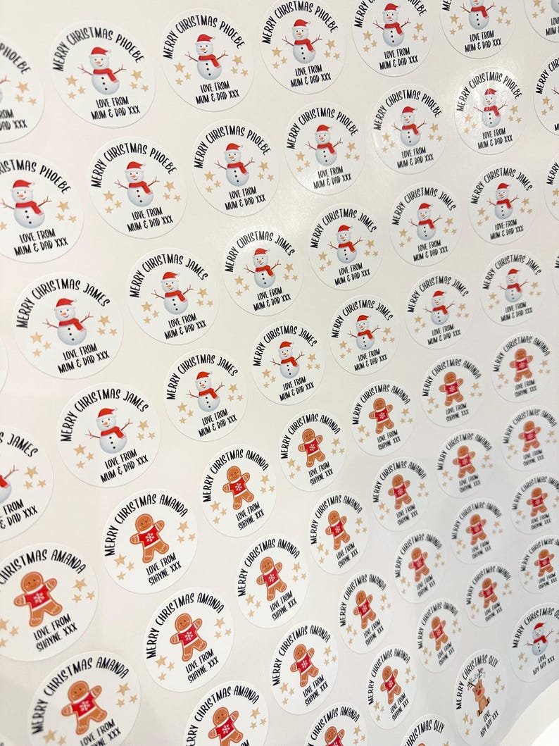 Op de afbeelding: Een vel kerststickers. De stickers tonen een sneeuwpop met een rode hoed en sjaal, en een peperkoekmannetje met een rode sjaal. Tekst bevat "Vrolijk Kerstfeest" en namen als "Phoebe" en "James".