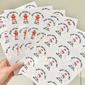 Op de afbeelding: Vellen witte kerststickers met diverse ontwerpen. Sommige stickers tonen een peperkoekmannetje, andere een sneeuwpop, en alle bevatten de tekst "Merry Christmas" en gepersonaliseerde namen. De stickers zijn rond en hebben gouden steraccenten.