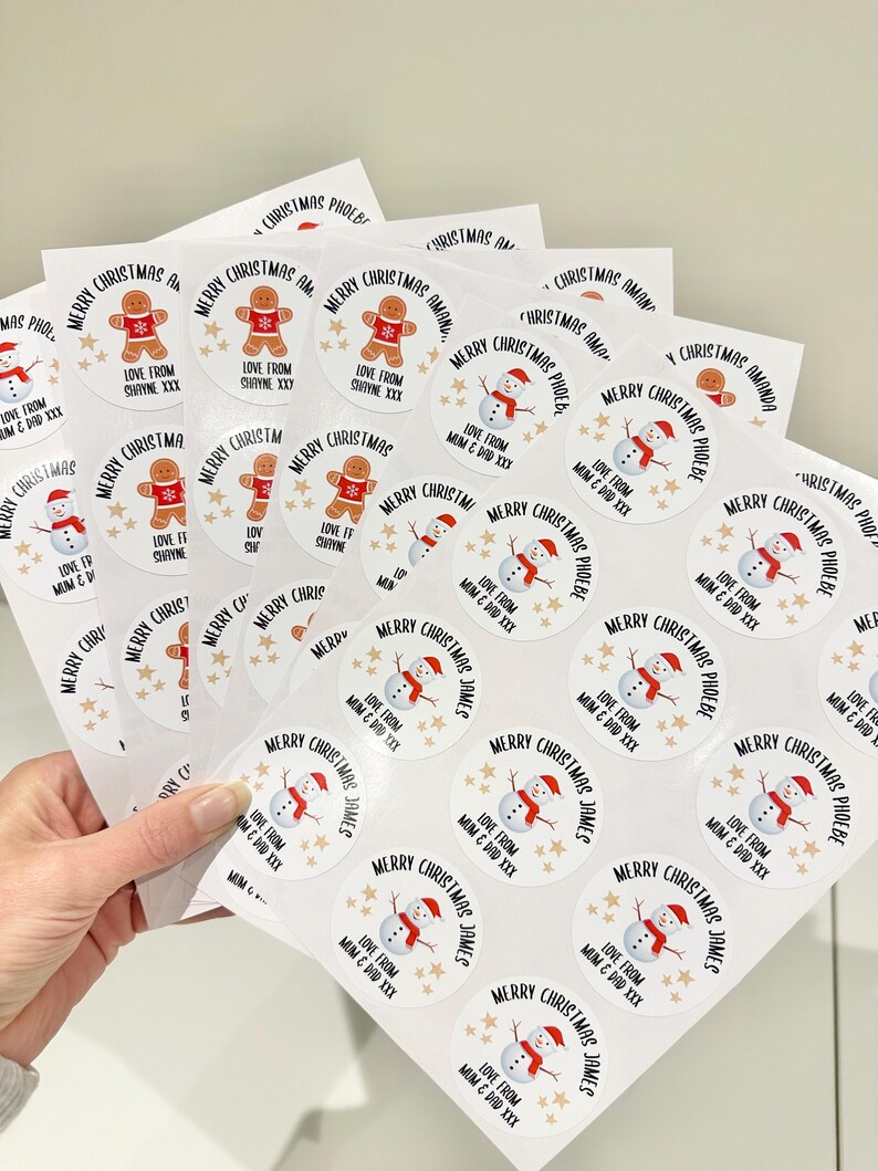 Op de afbeelding: Vellen witte kerststickers met diverse ontwerpen. Sommige stickers tonen een peperkoekmannetje, andere een sneeuwpop, en alle bevatten de tekst "Merry Christmas" en gepersonaliseerde namen. De stickers zijn rond en hebben gouden steraccenten.
