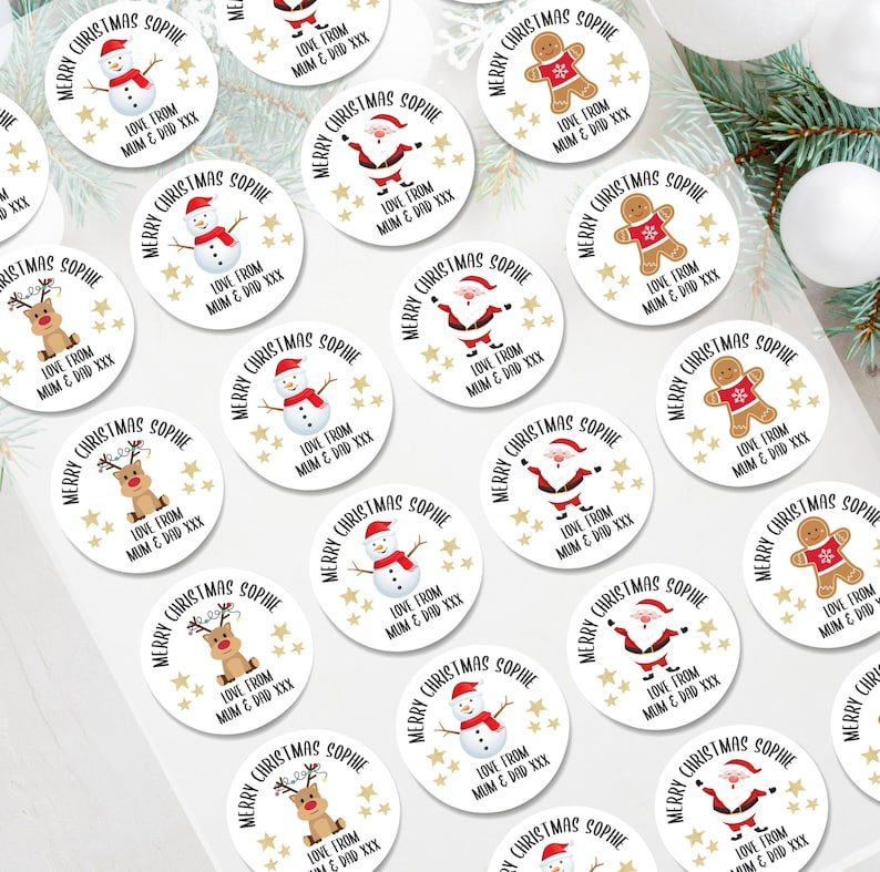 Op de afbeelding: Een verzameling witte, ronde kerststickers met diverse feestelijke illustraties, waaronder sneeuwmannen, peperkoekenmannen, rendieren en de kerstman. Elke sticker heeft de tekst "Merry Christmas Sophie" en "Love from Mum & Dad XXX".