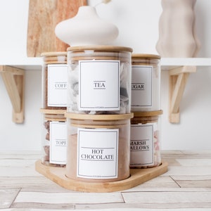 K&ouml;nnte beinhalten: Ein Set aus sechs K&uuml;chengl&auml;sern mit Bambusdeckeln, angeordnet auf einem Holztablett. Jedes Glas hat ein wei&szlig;es Etikett mit schwarzem Text, der den Inhalt wie "TEA" und "HOT CHOCOLATE" angibt. Die Gl&auml;ser sind mit verschiedenen Trockenwaren gef&uuml;llt.