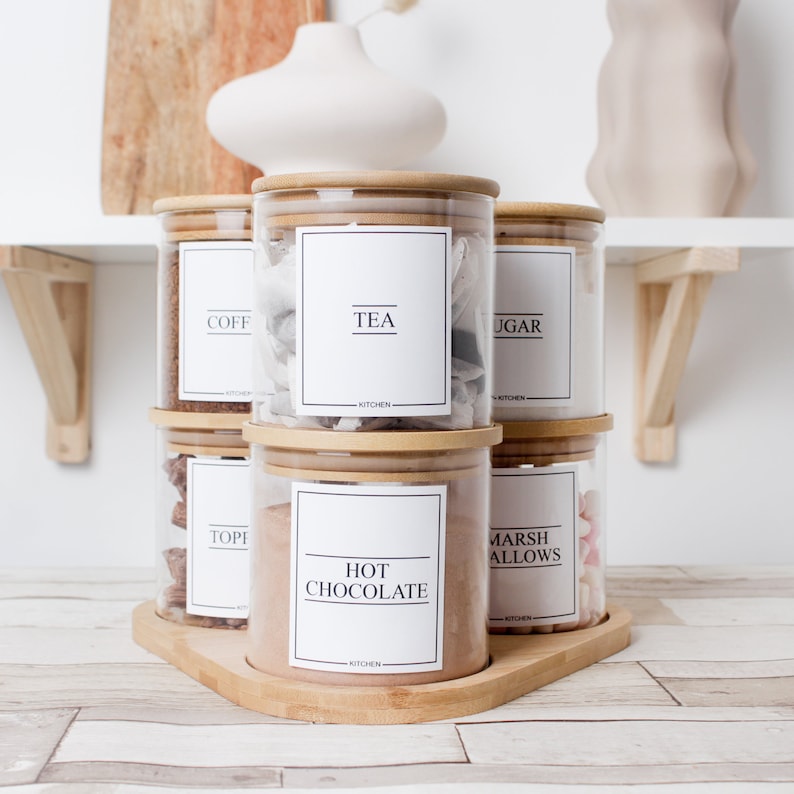 K&ouml;nnte beinhalten: Ein Set aus sechs K&uuml;chengl&auml;sern mit Bambusdeckeln, angeordnet auf einem Holztablett. Jedes Glas hat ein wei&szlig;es Etikett mit schwarzem Text, der den Inhalt wie "TEA" und "HOT CHOCOLATE" angibt. Die Gl&auml;ser sind mit verschiedenen Trockenwaren gef&uuml;llt.
