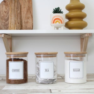 May include: Three glass kitchen canisters with bamboo lids labeled "COFFEE", "TEA", and "SUGAR". The coffee canister is filled with brown granules, the tea canister with tea bags, and the sugar canister with white sugar. A white shelf holds the canisters, a cutting board, and decorative items.