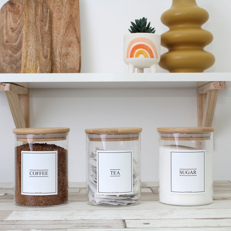 May include: Three glass kitchen canisters with bamboo lids labeled "COFFEE", "TEA", and "SUGAR". The coffee canister is filled with brown granules, the tea canister with tea bags, and the sugar canister with white sugar. A white shelf holds the canisters, a cutting board, and decorative items.