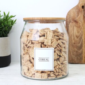 K&ouml;nnte beinhalten: Ein klares Glasgef&auml;&szlig; gef&uuml;llt mit M&uuml;sli, mit einem Bambusdeckel versehen. Ein wei&szlig;es Etikett auf der Vorderseite tr&auml;gt den schwarzen Text "CEREAL". Das Glas steht auf einer wei&szlig;en Oberfl&auml;che, mit einer Pflanze und einem Holzschneidebrett im Hintergrund.