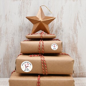 Op de afbeelding: Een stapel ingepakte cadeaus met rood en wit touw, bekroond met een gouden ster. De cadeaus zijn versierd met ronde stickers met vakantiegraphics en de tekst "Merry Christmas Sophie" en "Love From Mom & Dad XXX".