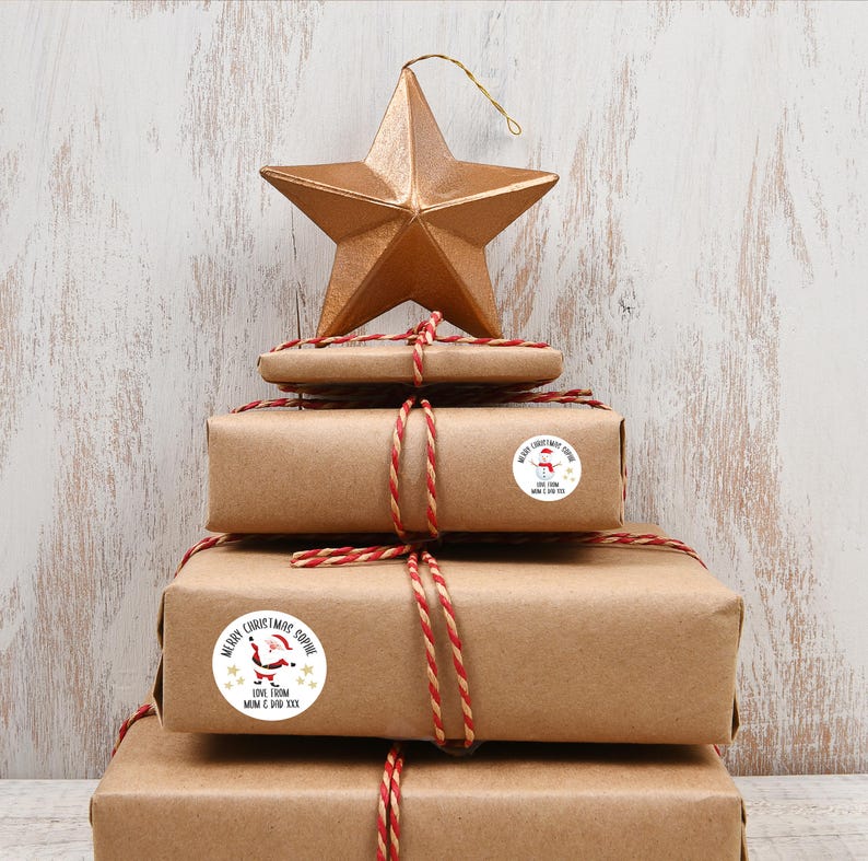 Op de afbeelding: Een stapel ingepakte cadeaus met rood en wit touw, bekroond met een gouden ster. De cadeaus zijn versierd met ronde stickers met vakantiegraphics en de tekst "Merry Christmas Sophie" en "Love From Mom & Dad XXX".