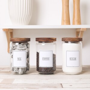 K&ouml;nnte beinhalten: Drei transparente Glasbeh&auml;lter mit Holzdeckeln, beschriftet mit "TEA", "COFFEE" und "SUGAR". Die Gl&auml;ser sind mit Teebeuteln, Kaffeebohnen und Zucker gef&uuml;llt. Sie stehen auf einer Holzoberfl&auml;che, mit einem wei&szlig;en Regal und dekorativen Elementen im Hintergrund.
