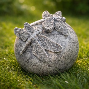 May include: A spherical gray stone garden ornament featuring two sculpted dragonflies with detailed wings resting on the top surface. The object has a weathered, textured finish and a rounded shape, showcasing intricate insect carvings.