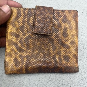 May include: A square-shaped bi-fold wallet featuring a textured snakeskin pattern in shades of dark brown and mustard yellow. The wallet includes a central vertical tab closure that secures the front flap, showcasing a consistent reptilian scale texture across the entire exterior surface of the accessory.