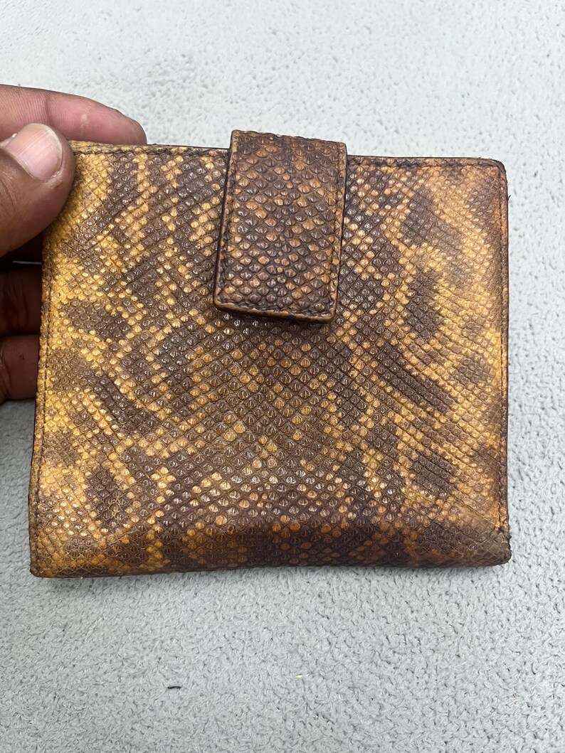May include: A square-shaped bi-fold wallet featuring a textured snakeskin pattern in shades of dark brown and mustard yellow. The wallet includes a central vertical tab closure that secures the front flap, showcasing a consistent reptilian scale texture across the entire exterior surface of the accessory.