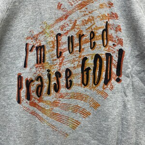 May include: A heather gray sweatshirt featuring a graphic print with the black text "I'm cured. praise GOD!" layered over a textured, distressed background of orange, brown, and yellow brushstrokes that form a heart-like shape on the front.