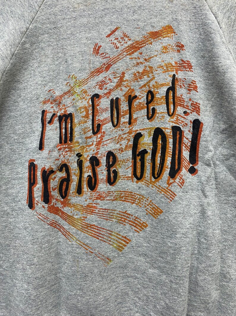 May include: A heather gray sweatshirt featuring a graphic print with the black text "I'm cured. praise GOD!" layered over a textured, distressed background of orange, brown, and yellow brushstrokes that form a heart-like shape on the front.