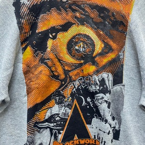 May include: A heather gray crewneck sweatshirt featuring a large graphic print on the front. The design displays a stylized orange and black eye, a figure in a bowler hat, and the text "CLOCKWORK ORANGE" in bold orange lettering at the bottom.