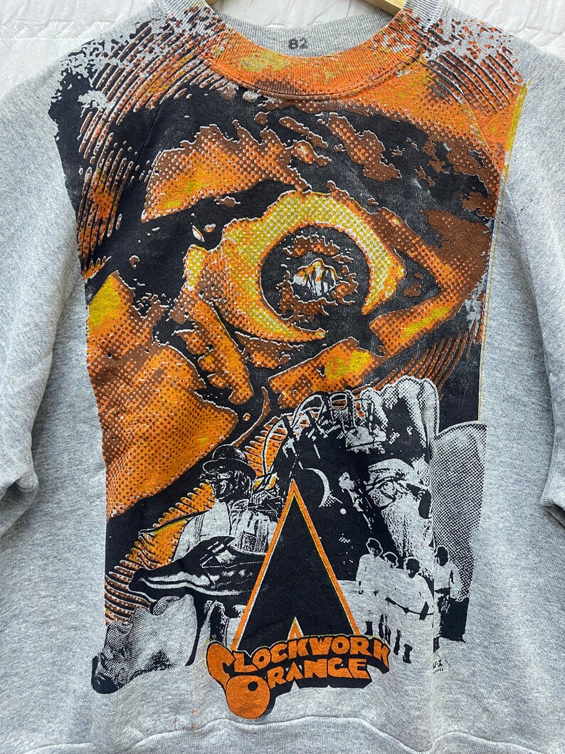May include: A heather gray crewneck sweatshirt featuring a large graphic print on the front. The design displays a stylized orange and black eye, a figure in a bowler hat, and the text "CLOCKWORK ORANGE" in bold orange lettering at the bottom.