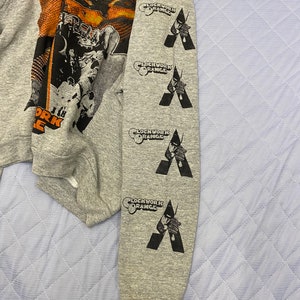 May include: A heather gray long-sleeve sweatshirt featuring a large graphic print on the chest and four repeating black vertical prints on the sleeve. The sleeve prints display the text Clockwork Orange above a stylized figure holding a cane.