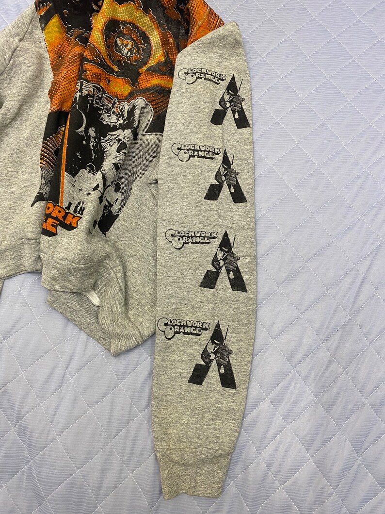 May include: A heather gray long-sleeve sweatshirt featuring a large graphic print on the chest and four repeating black vertical prints on the sleeve. The sleeve prints display the text Clockwork Orange above a stylized figure holding a cane.