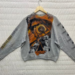 May include: A heather gray long-sleeve sweatshirt featuring a large, orange-toned graphic of a human eye on the front. The text "I'm cured! Praise God!" is printed on the right sleeve, while "Clockwork Orange" appears on the left sleeve and chest.