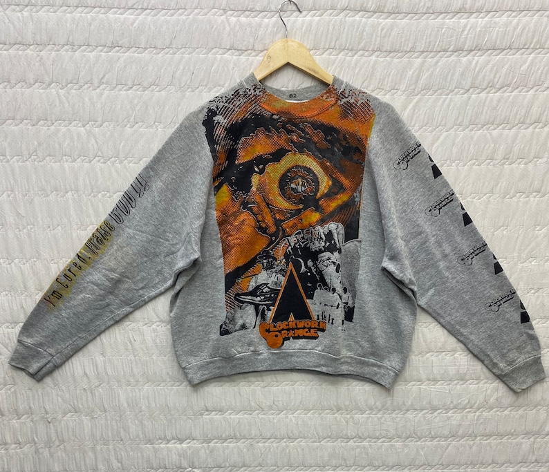 May include: A heather gray long-sleeve sweatshirt featuring a large, orange-toned graphic of a human eye on the front. The text "I'm cured! Praise God!" is printed on the right sleeve, while "Clockwork Orange" appears on the left sleeve and chest.