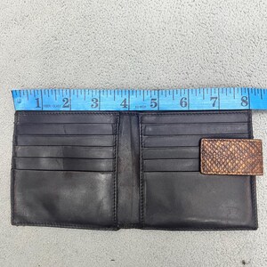 May include: A black leather bifold wallet with multiple card slots and a brown snakeskin-textured tab closure. A blue measuring tape is positioned along the top edge, indicating a width of approximately 8 inches for the open wallet.