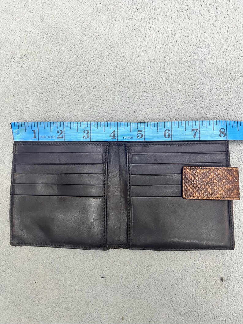 May include: A black leather bifold wallet with multiple card slots and a brown snakeskin-textured tab closure. A blue measuring tape is positioned along the top edge, indicating a width of approximately 8 inches for the open wallet.