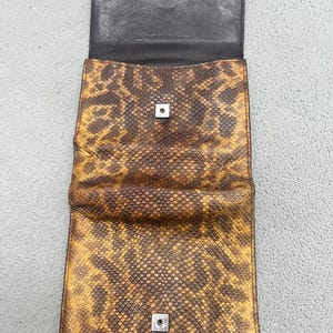 May include: A rectangular wallet featuring a brown and yellow snakeskin pattern. The wallet is open, revealing a dark brown interior lining and two silver-toned metal snap button closures positioned on the flap and the main body of the wallet.