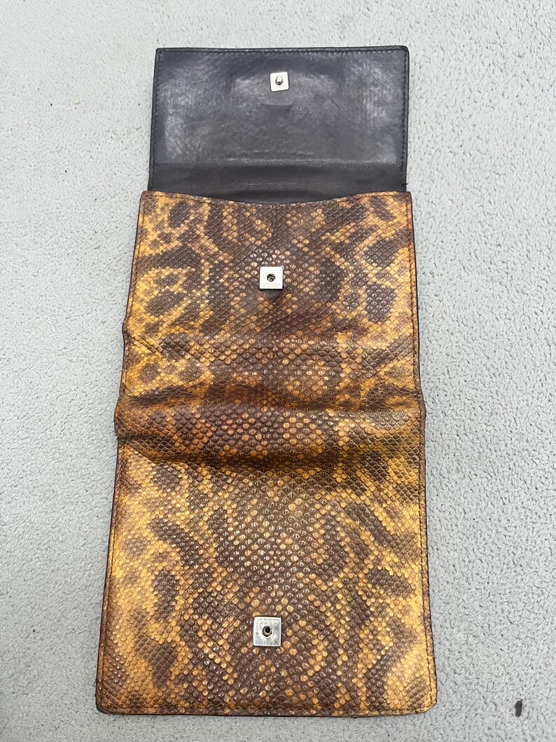 May include: A rectangular wallet featuring a brown and yellow snakeskin pattern. The wallet is open, revealing a dark brown interior lining and two silver-toned metal snap button closures positioned on the flap and the main body of the wallet.
