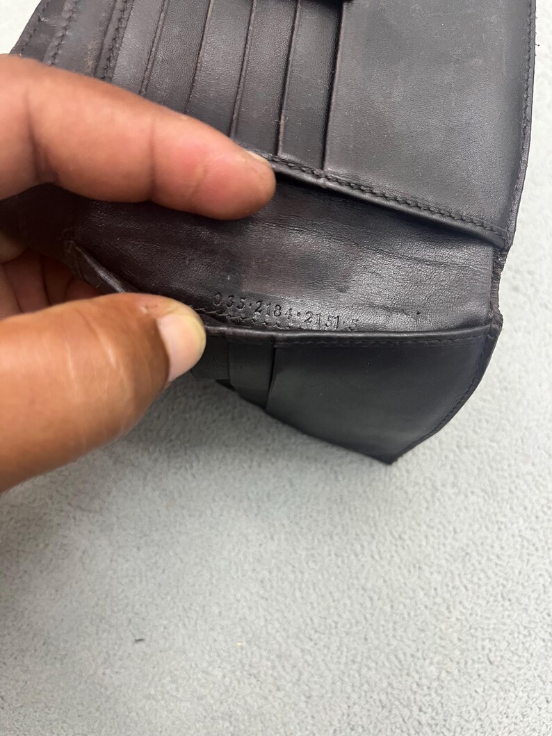 May include: A dark brown leather wallet featuring multiple interior card slots and a visible serial number embossed on the inner fold reading 035 2184 2151 5. The wallet has a smooth texture and a structured design with visible stitching along the edges.