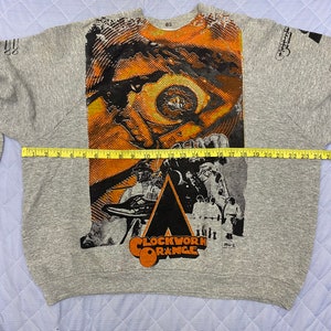 May include: A heather gray sweatshirt featuring a large graphic print from A Clockwork Orange, including a stylized eye and the film title. A yellow measuring tape is placed across the chest, showing a width of approximately 24 inches.