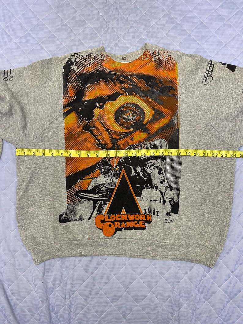 May include: A heather gray sweatshirt featuring a large graphic print from A Clockwork Orange, including a stylized eye and the film title. A yellow measuring tape is placed across the chest, showing a width of approximately 24 inches.