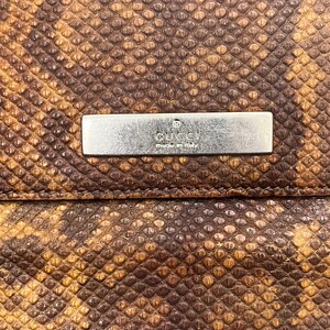 May include: A brown and tan snakeskin-textured wallet featuring a rectangular silver-toned metal plaque centered on the front. The plaque is engraved with the text GUCCI and made in italy, with a small registered trademark symbol above the brand name.