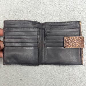 May include: A bi-fold wallet featuring a dark charcoal interior with multiple horizontal card slots on both sides. The exterior flap is crafted from a textured, snakeskin-patterned material in shades of brown and tan, secured with a snap closure.
