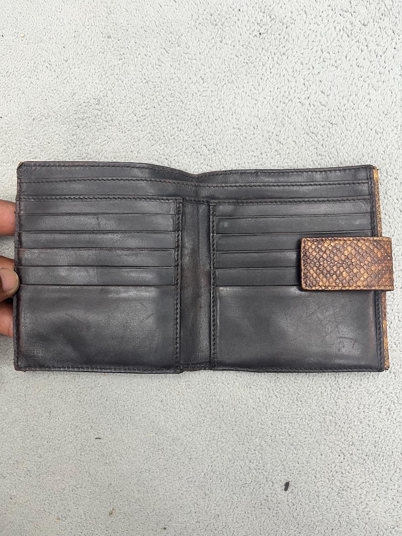 May include: A bi-fold wallet featuring a dark charcoal interior with multiple horizontal card slots on both sides. The exterior flap is crafted from a textured, snakeskin-patterned material in shades of brown and tan, secured with a snap closure.