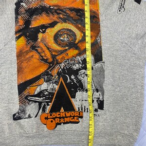 May include: A heather gray sweatshirt featuring a large graphic print of the movie Clockwork Orange, including a close-up of an eye and characters in white outfits. A yellow measuring tape is placed vertically, showing a length of 25 inches.