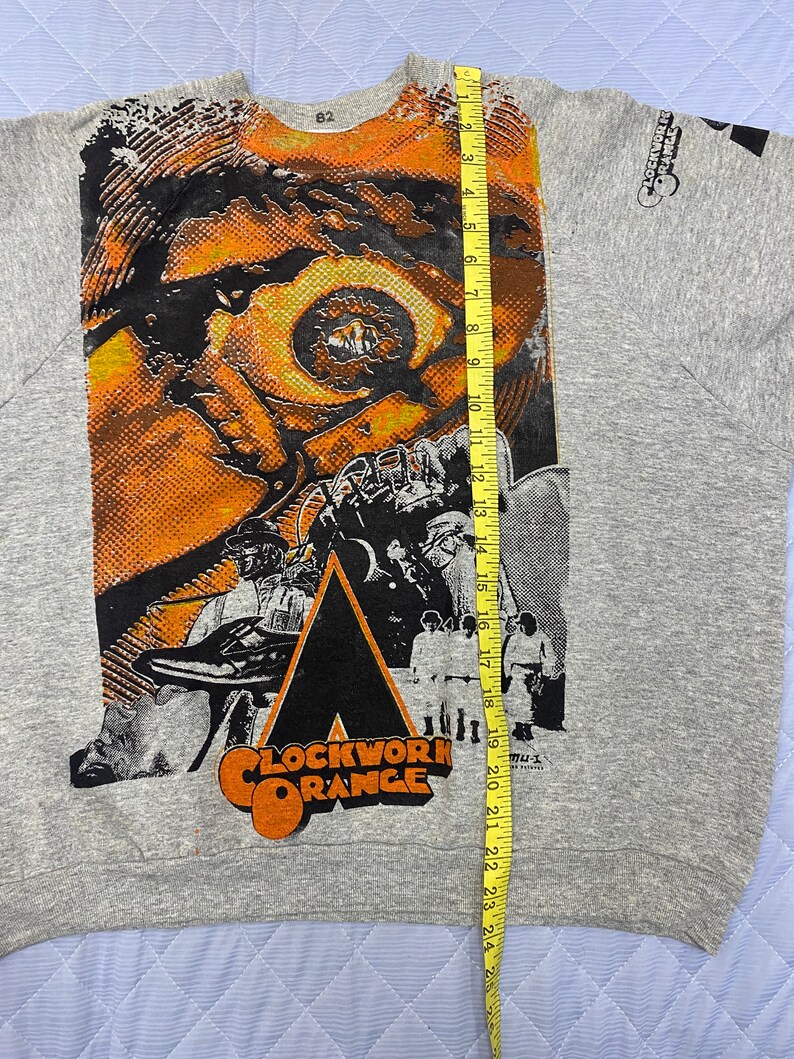May include: A heather gray sweatshirt featuring a large graphic print of the movie Clockwork Orange, including a close-up of an eye and characters in white outfits. A yellow measuring tape is placed vertically, showing a length of 25 inches.