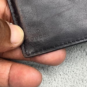 May include: A black leather wallet featuring a smooth, textured surface with visible stitching along the edge. The word GUCCI is embossed into the leather material, accompanied by a small circular symbol positioned directly above the brand name.