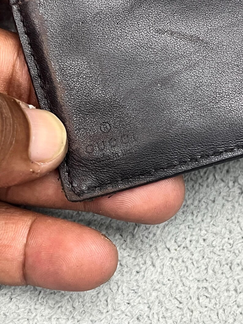 May include: A black leather wallet featuring a smooth, textured surface with visible stitching along the edge. The word GUCCI is embossed into the leather material, accompanied by a small circular symbol positioned directly above the brand name.
